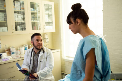 A transgender woman in a hospital gown speaking to her doctor, a transgender man, in an exam room