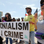 Individuals holding a sign that says Colonialism is a Crime.